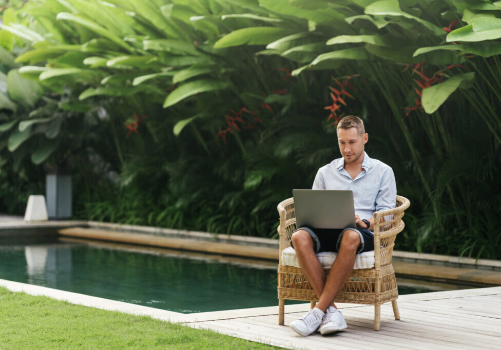 Young man using a laptop computer in a garden with a swimming pool. business, study, freelance